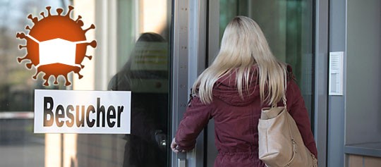 Eine Frau steht vor einer Glastür mit der Aufschrift "Besucher", daneben die Illustration eines Virus mit Mundschutz