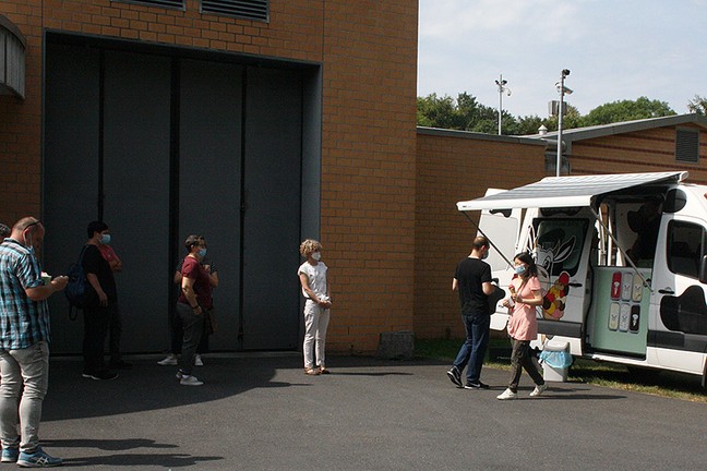 Mehrere Personen stehen mit räumlichem Abstand vor einem Eiswagen an. Der Eiswagen steht vor einer Klinikmauer.