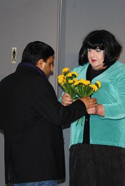 Ein Mann und ein als Frau kostümierter Mann tauschen vor einer Herrentoilette Blumen aus